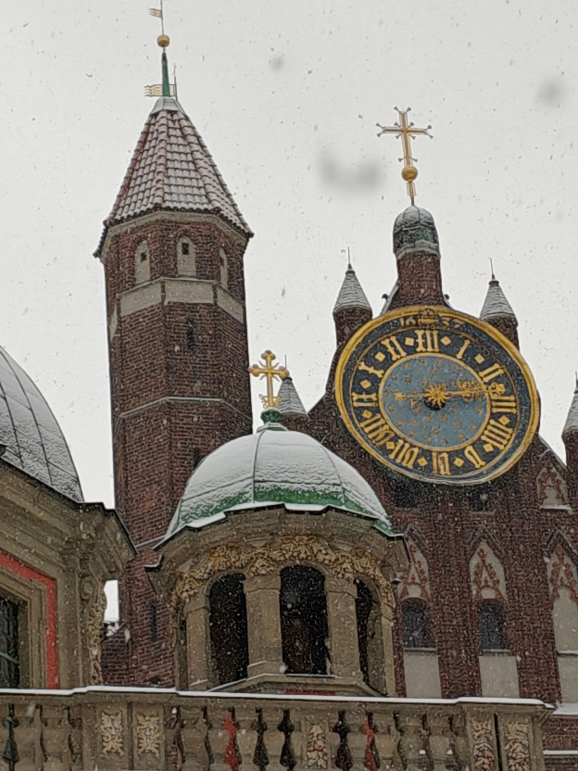 Bakstenen gebouw met toren, klok en gouden kruisen; het sneeuwt.