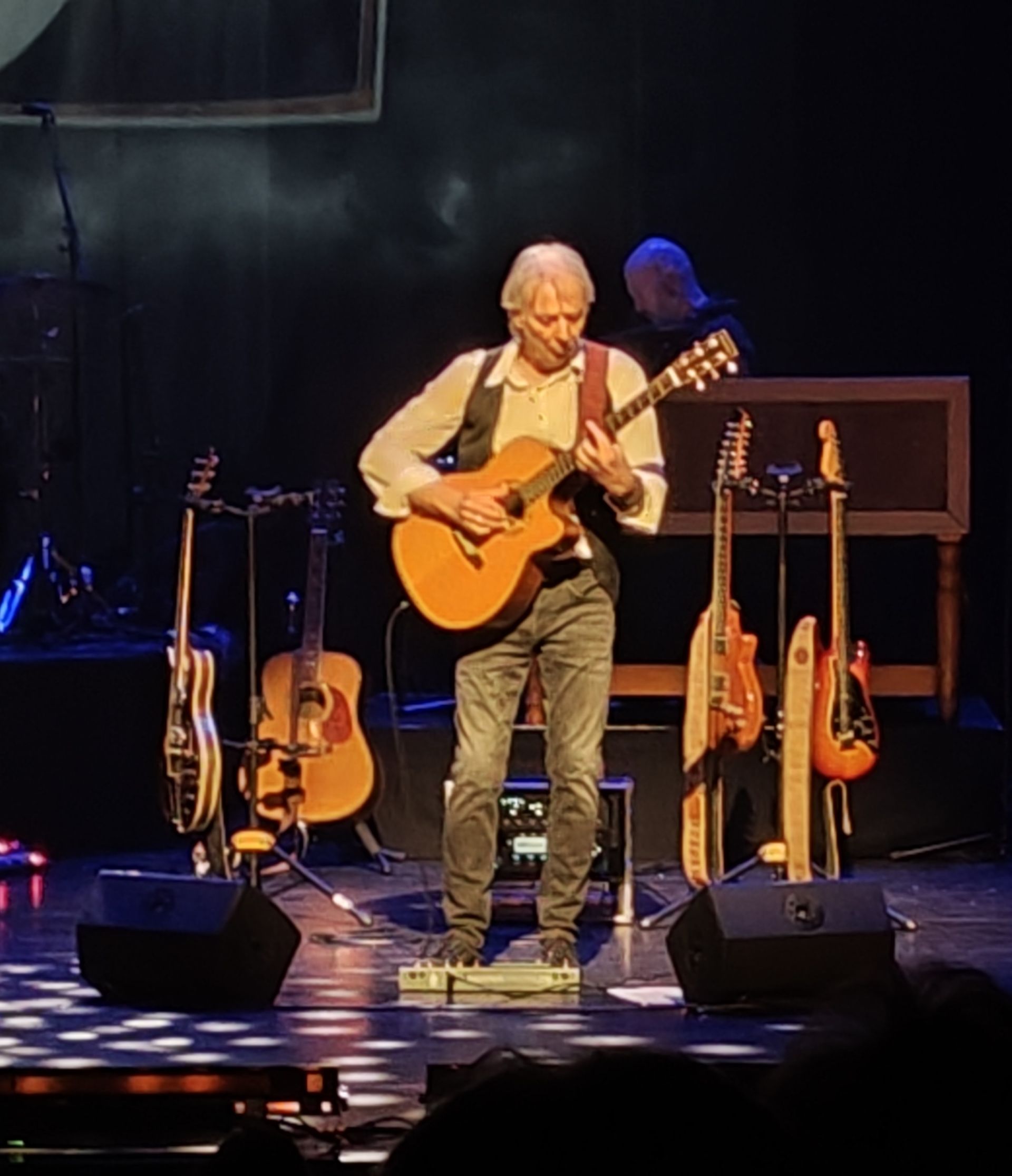 Muzikant op het podium die akoestische gitaar speelt. Gitaren op de achtergrond, donkere omgeving.