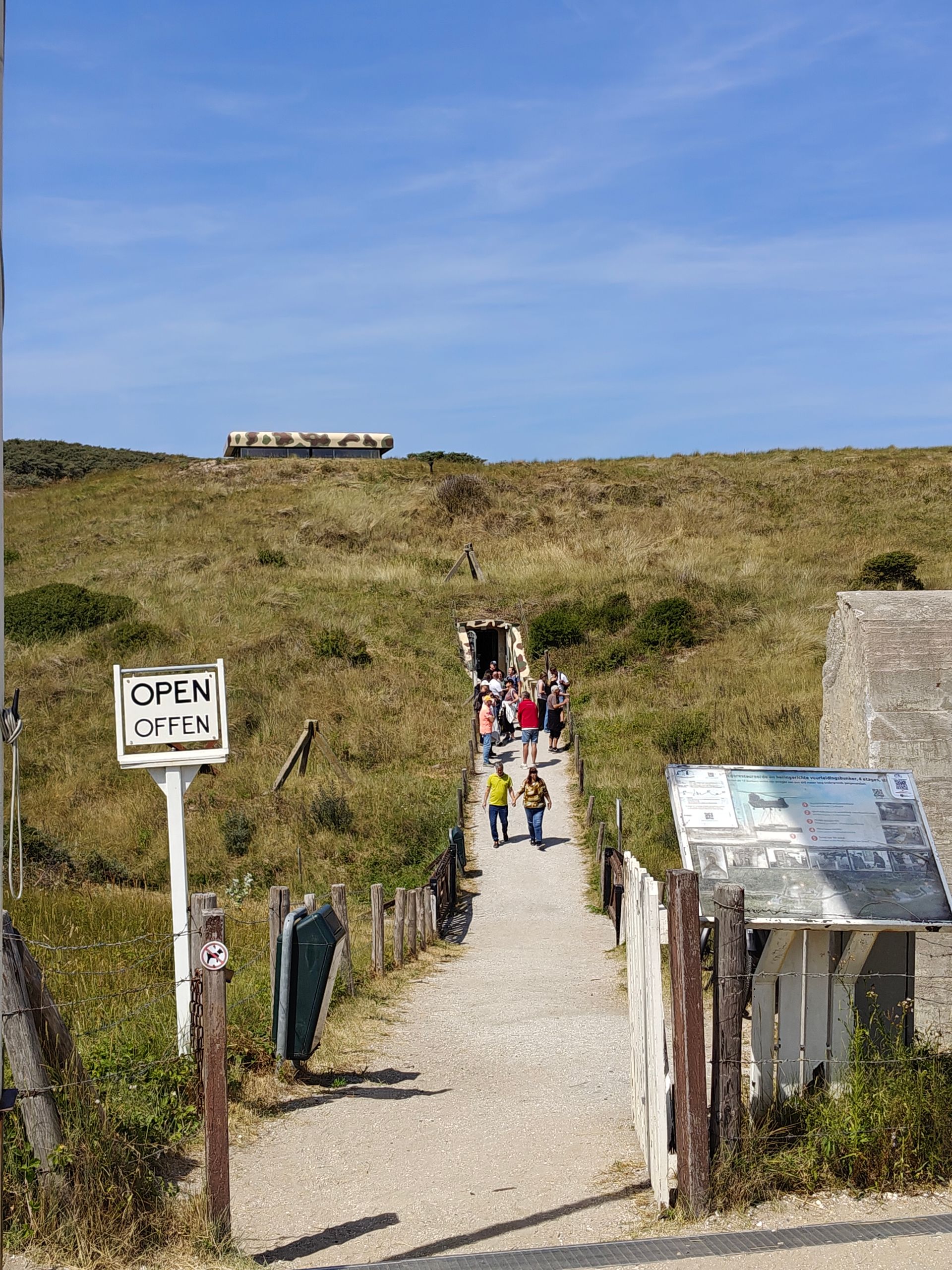 Een zandpad door de duinen leidt naar een strand, waar verschillende mensen onder een blauwe hemel wandelen. Open