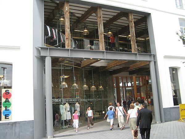 Mensen betreden een modern gebouw met een glazen gevel, hangende lampen en een kleurrijk verticaal reclamebord.