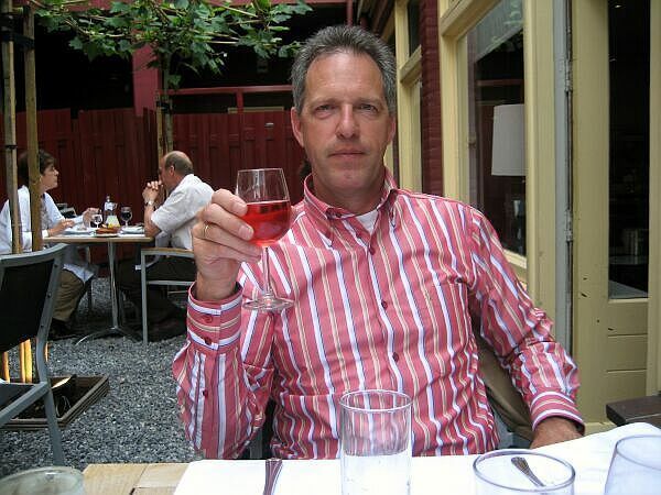 Een man in een roze gestreept shirt heft zijn glas aan een tafeltje op een terras.