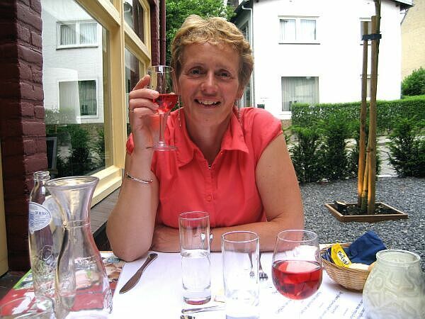 Vrouw in koraalkleurig shirt lacht aan een tafeltje op een terrasje met drankjes en een vaas.