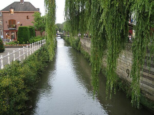 Een smal kanaal tussen stenen muren en huizen, met overhangende wilgen en kalm water.