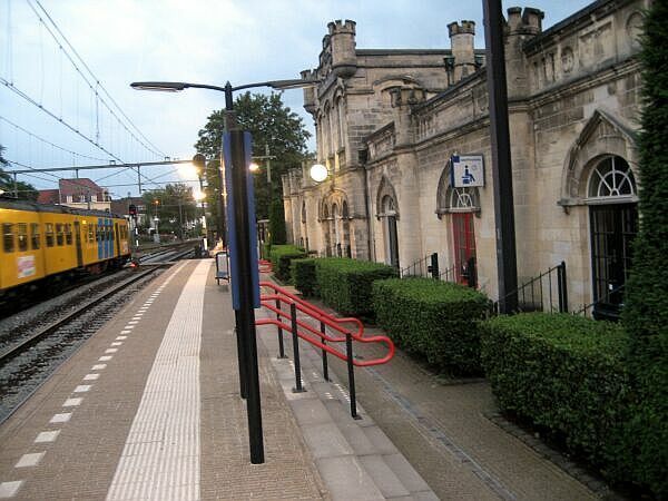 Perron naast een stenen stationsgebouw, met een gele tram en rode leuningen langs het voetpad.