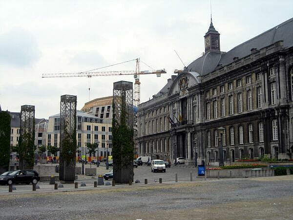 Stadsplein met een historisch stenen gebouw, moderne torens en een bouwkraan onder een bewolkte hemel.