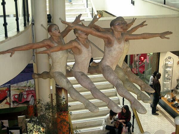 Drie bronzen figuren strekken zich uit terwijl ze een trap afdalen in een lichte, overdekte atrium.