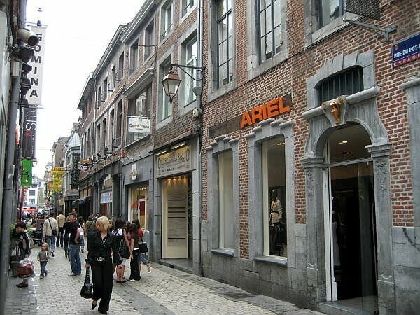 Smalle bakstenen winkelstraat met voetgangers en winkelpanden, waaronder een oranje verlicht Ariel-reclamebord.