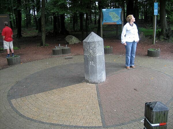 Een bospad met een centrale stenen markering en twee mensen die er vlakbij staan ​​op een rond, geplaveid plein.