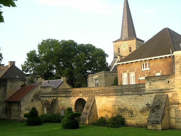 Een stenen muur en een boogbrug naast een kerk met een hoge torenspits en bomen onder een heldere hemel.