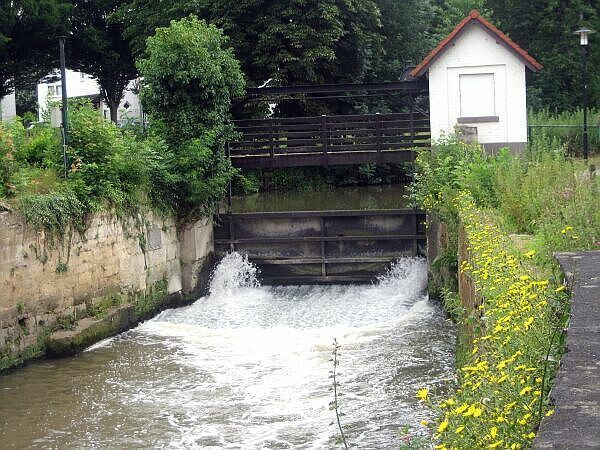 Een kleine waterval onder een brug, omgeven door groen en gele wilde bloemen, met een wit schuurtje in de buurt.
