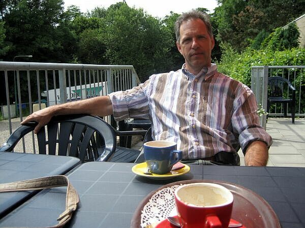 Een man zit aan een tafeltje op een terras met een kop koffie en een blauwe mok, omgeven door groen.