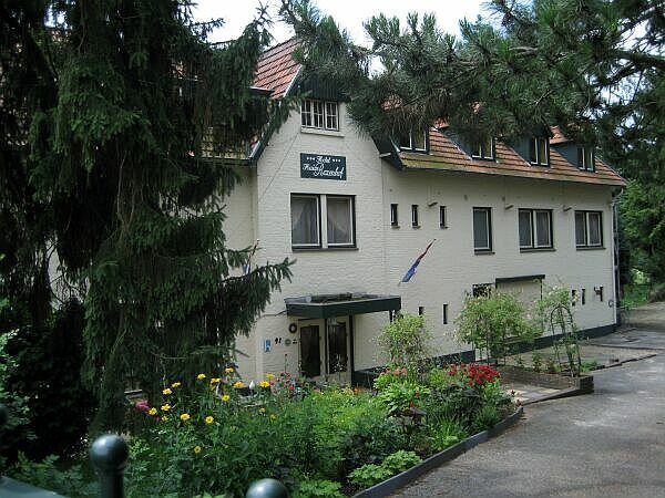 Wit huis met rood dak naast een tuin en oprit, omgeven door hoge bomen.