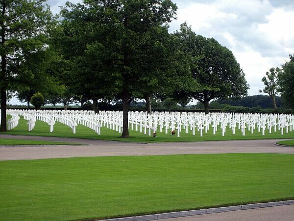 Rijen witte kruisen op een begraafplaats onder grote bomen op een groen gazon.