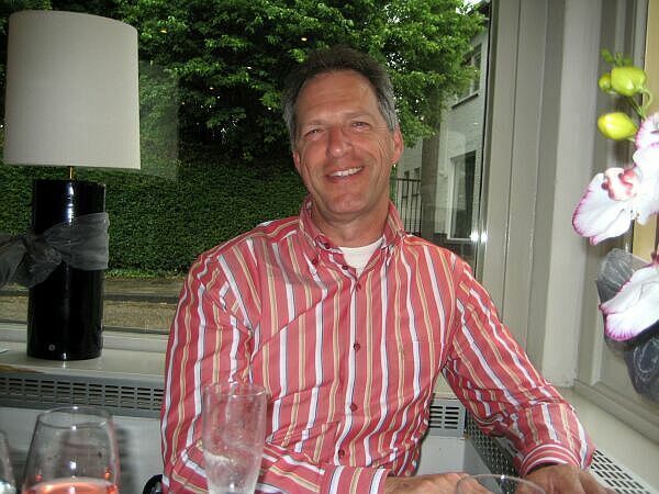 Een lachende man in een roze gestreept shirt zit aan een tafeltje op een terras met bloemen en een lamp in de buurt.