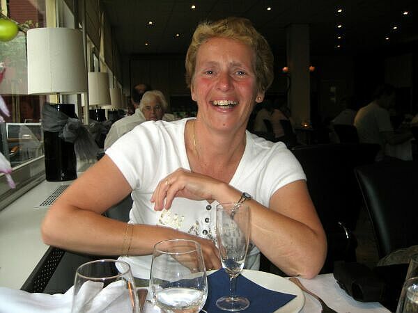 Een lachende vrouw in een witte blouse zit aan een restauranttafel met een wijnglas.