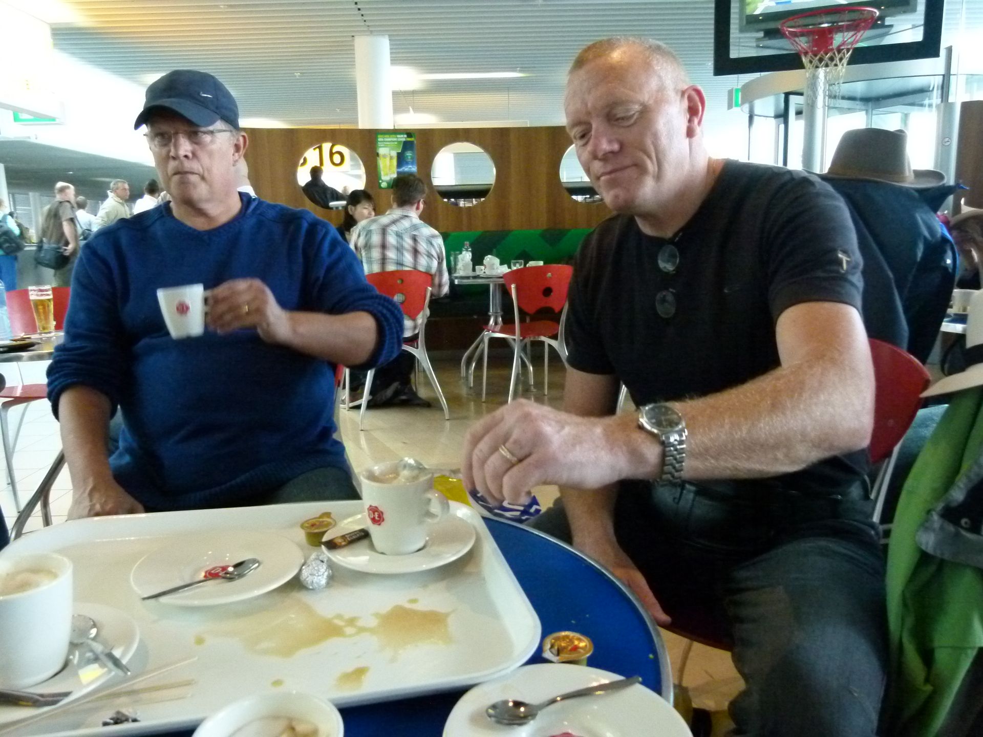Twee mannen zitten aan een tafeltje in een café en drinken koffie. De ene draagt ​​een blauwe pet, de andere een zwart shirt. De scène speelt zich af op een vliegveld.