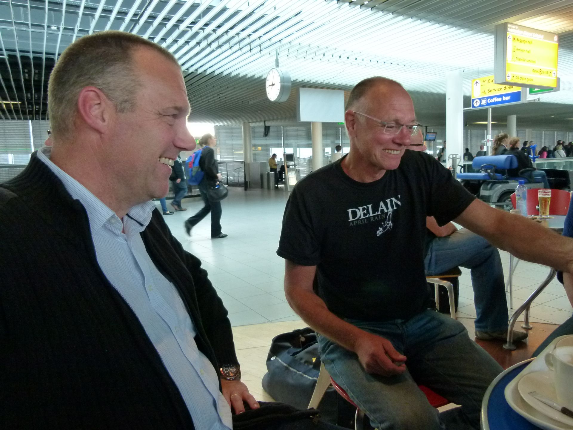 Twee mannen glimlachen aan een tafeltje in een café op een vliegveld. De ene draagt ​​een zwart shirt, de andere een jas.