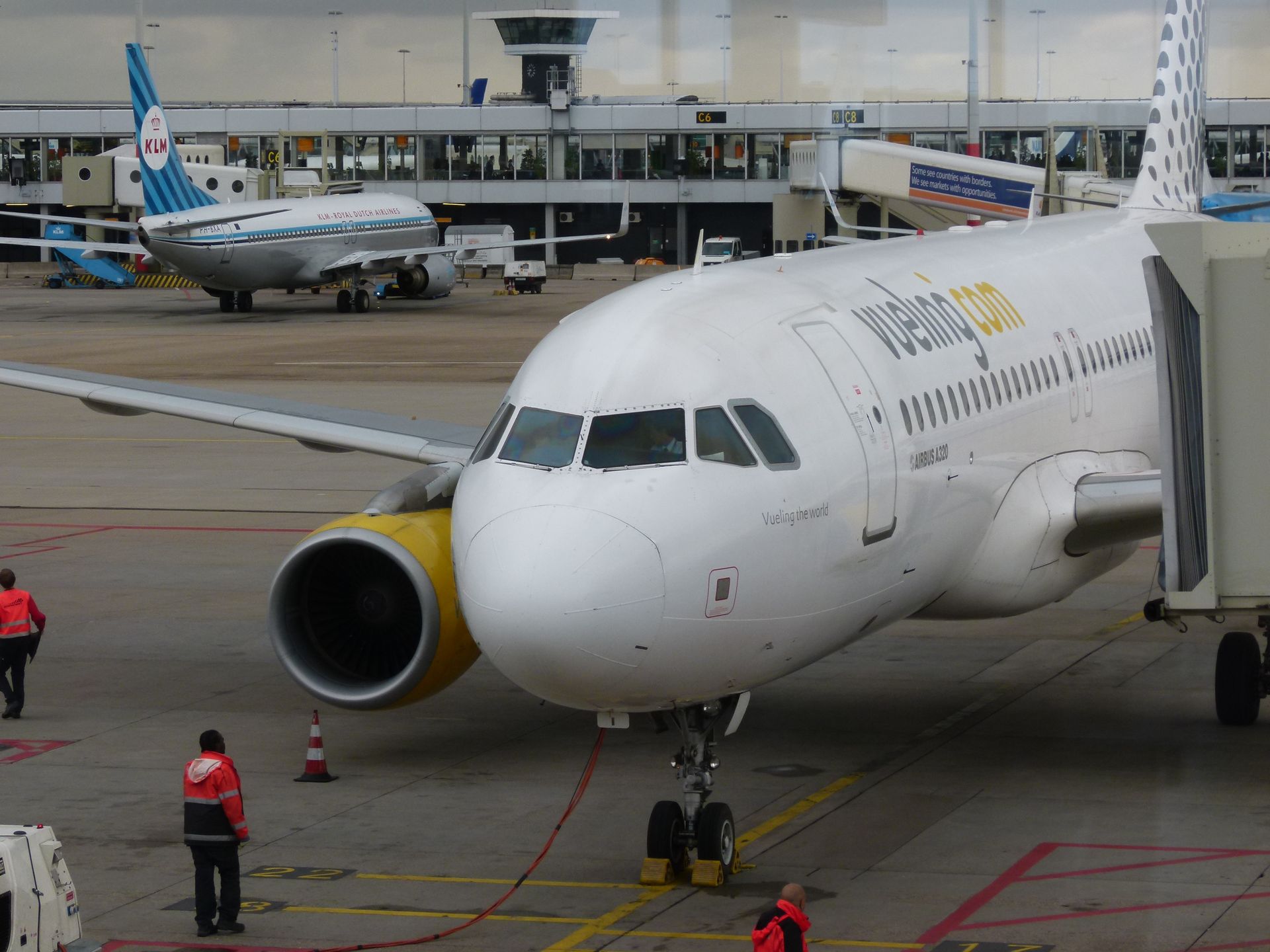 Wit Vueling-vliegtuig bij de gate, gele motordetails zichtbaar, mensen en de verkeerstoren op de achtergrond.