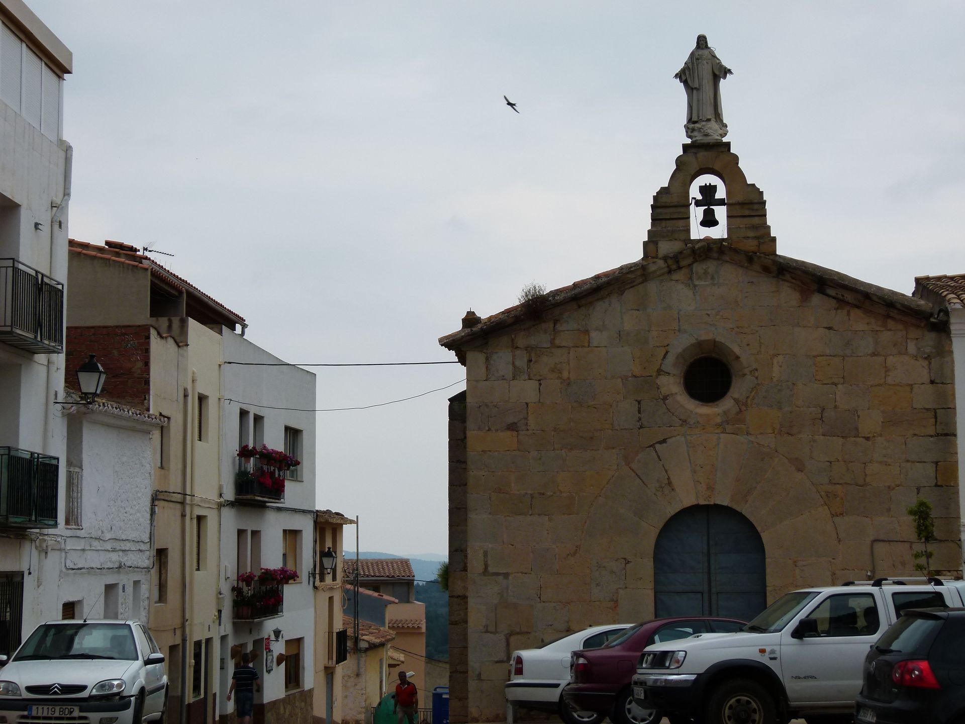Stenen kerk met klokkentoren en standbeeld, met uitzicht op een straat met geparkeerde auto's en gebouwen.