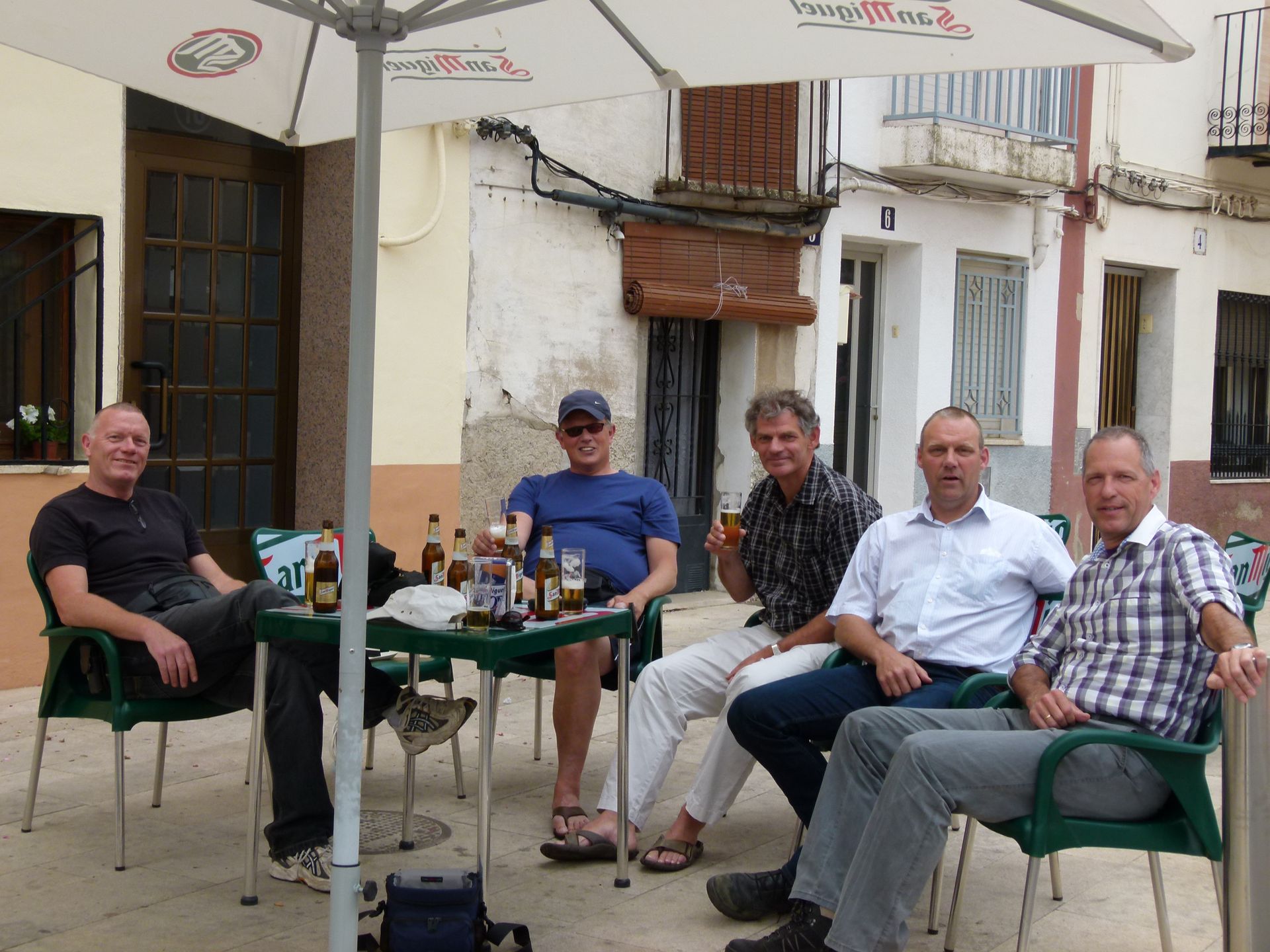 Vijf mannen zitten aan een tafeltje buiten en drinken bier onder een parasol op een Europees stadsplein.