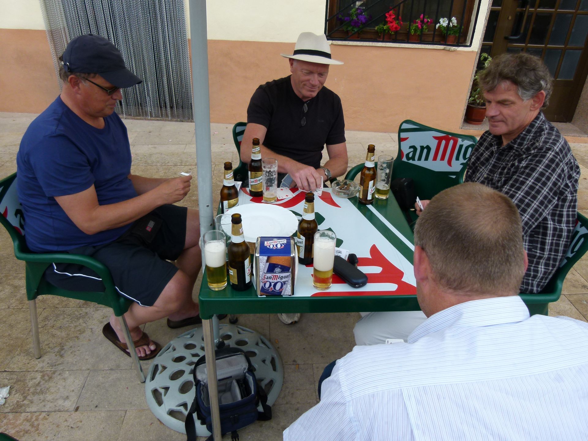 Vier mannen zitten buiten rond een tafel met biertjes. Een van hen steekt een aansteker aan. Groen-witte tafel, beige achtergrond.