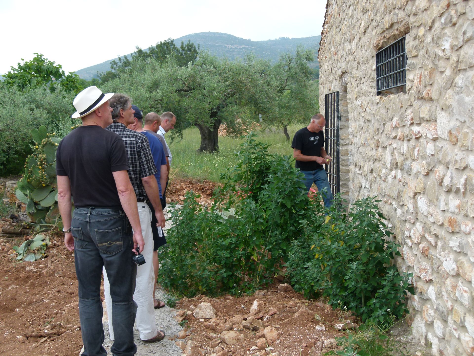 Een groep mannen inspecteert een stenen gebouw met een klein raam. Op de achtergrond zijn struiken en bomen te zien.