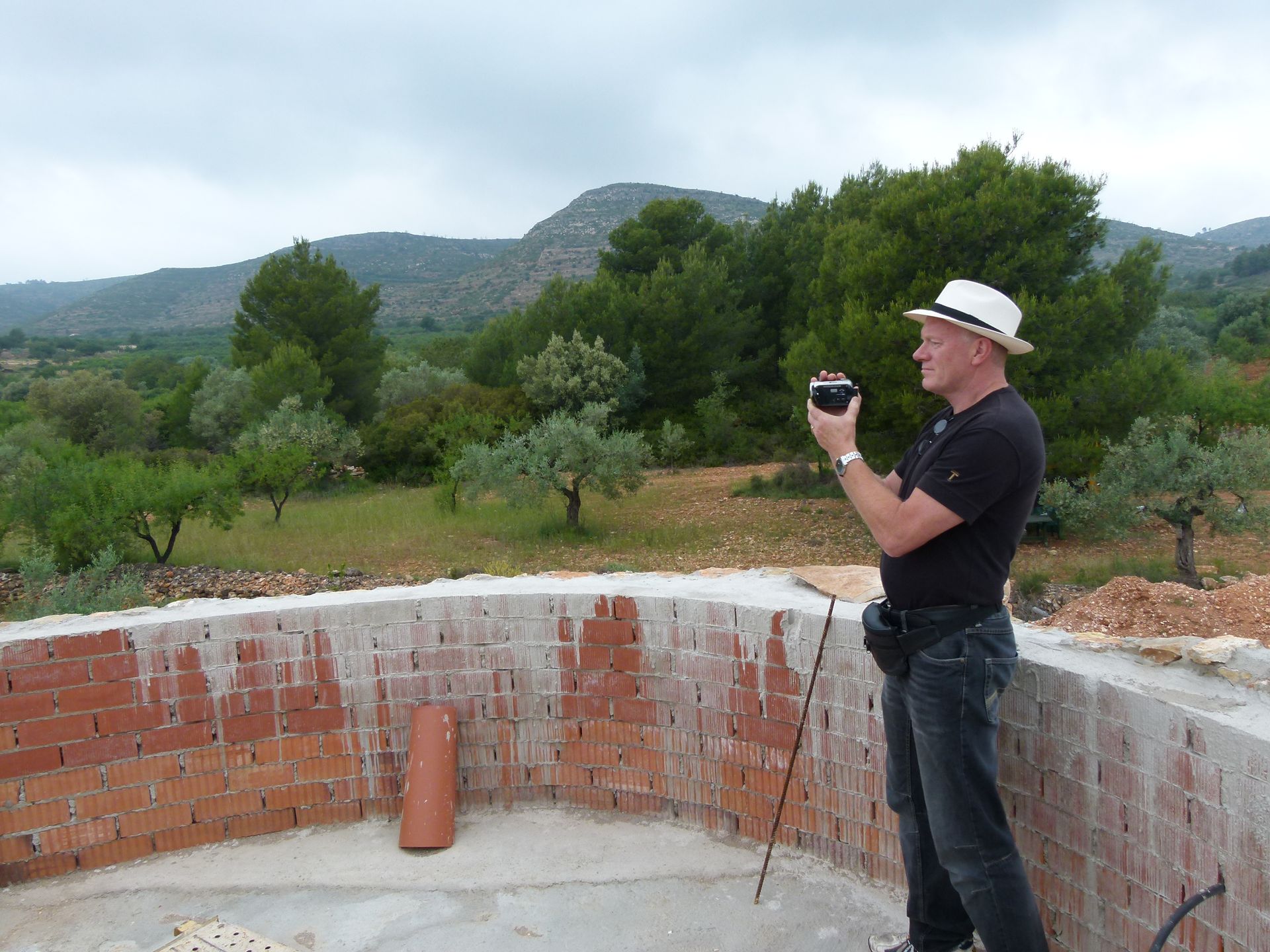 Een man met een hoed fotografeert een schilderachtig landschap vanaf een bakstenen gebouw. ​​Bergen en bomen zijn zichtbaar op de achtergrond.