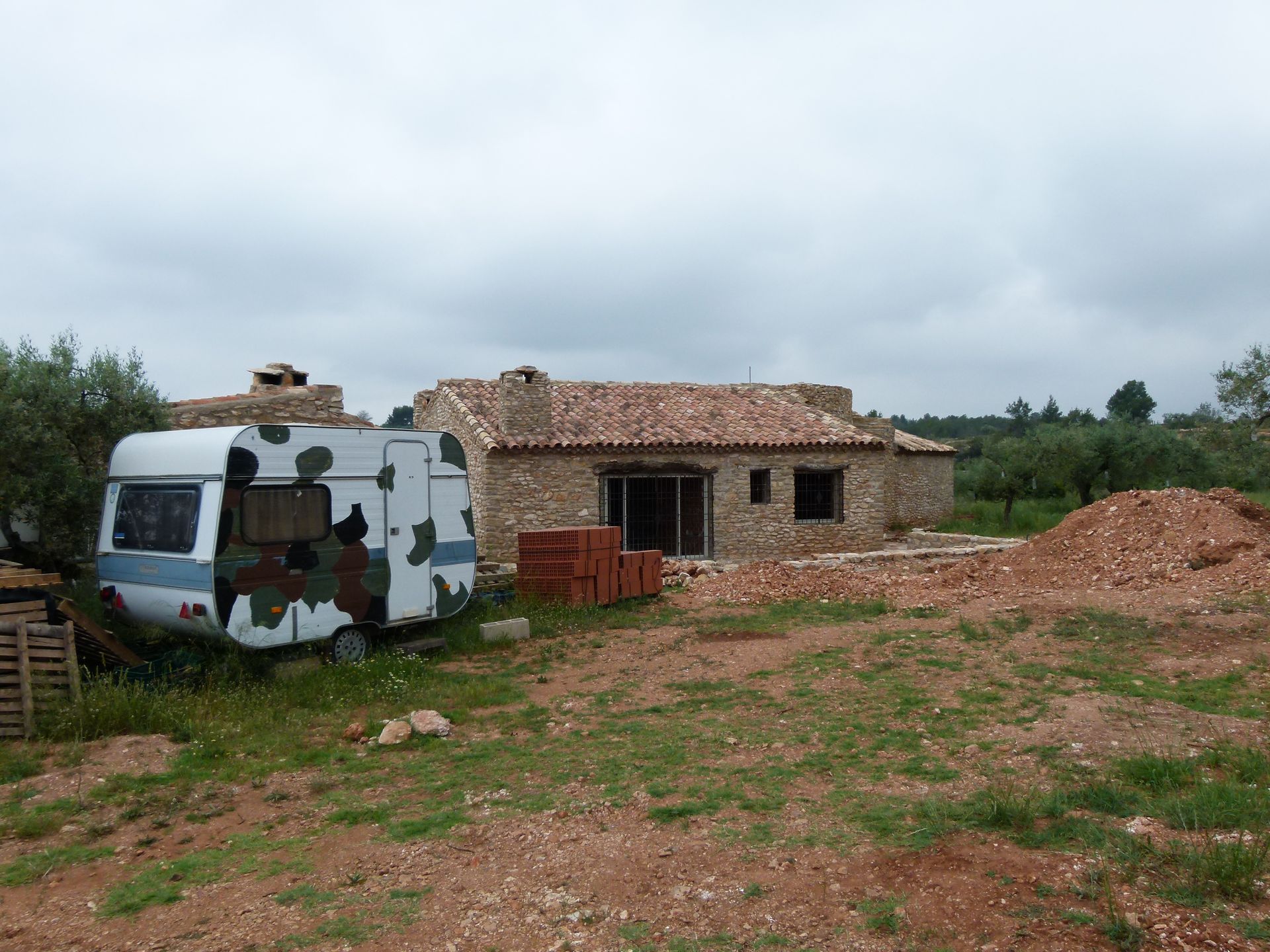 Een stenen huis in aanbouw met een gecamoufleerde camper op de voorgrond op een bewolkte dag.