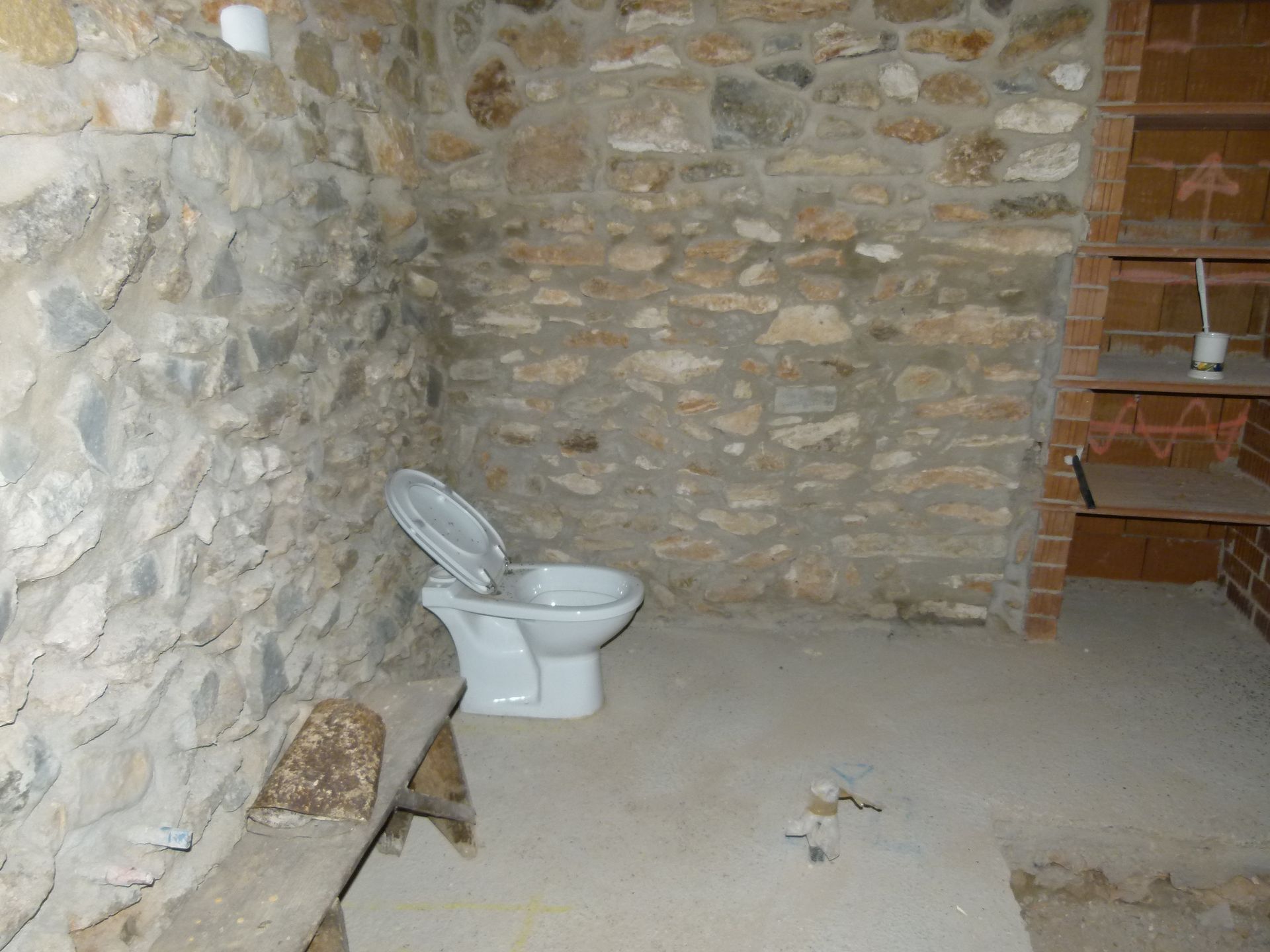 Toilet in een rustieke ruimte met stenen muren. Betonnen vloer met een houten bankje links en planken rechts.