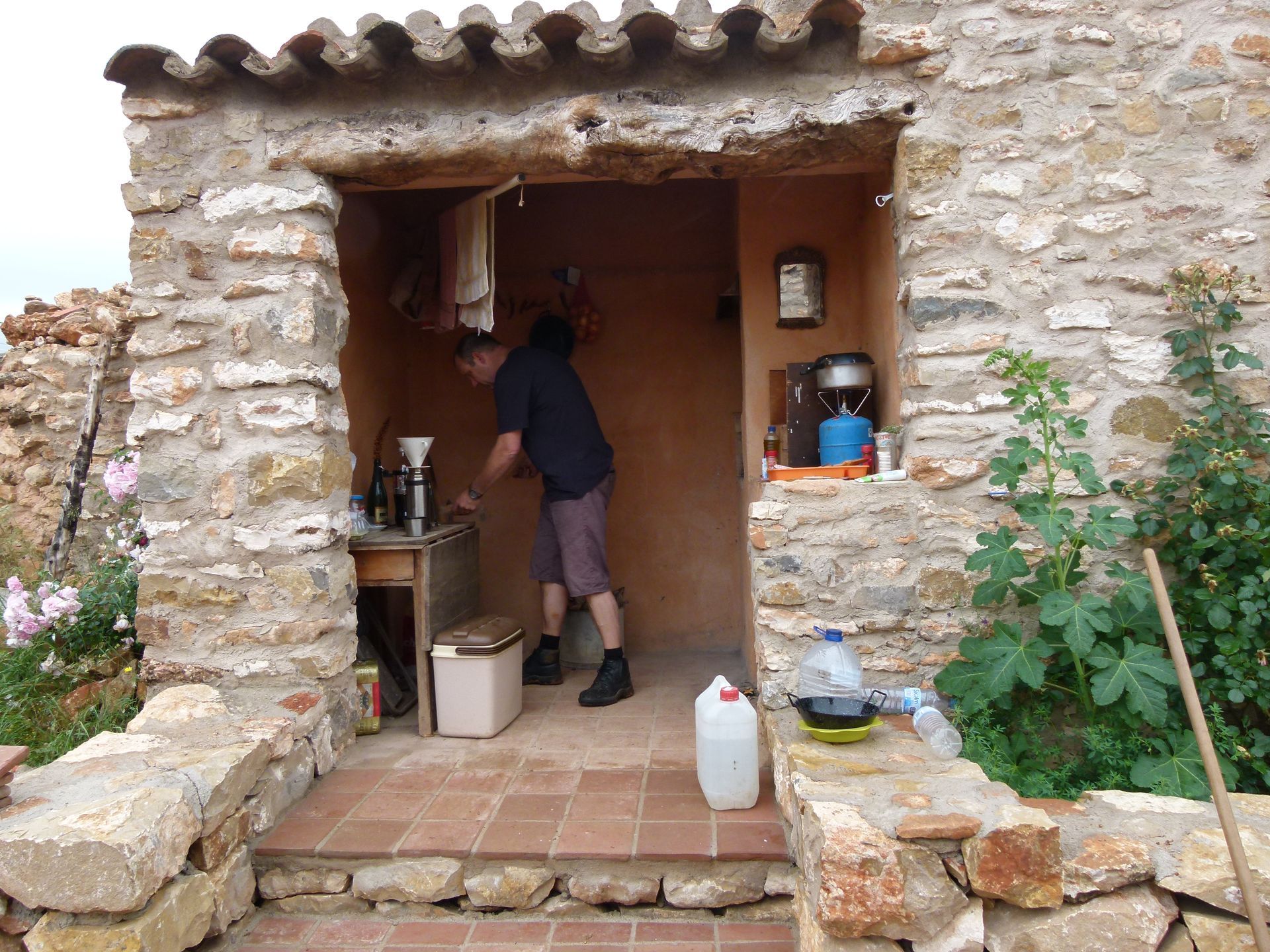 Persoon in de deuropening van een stenen gebouw, kookopstelling zichtbaar. Trap leidt naar de ingang.