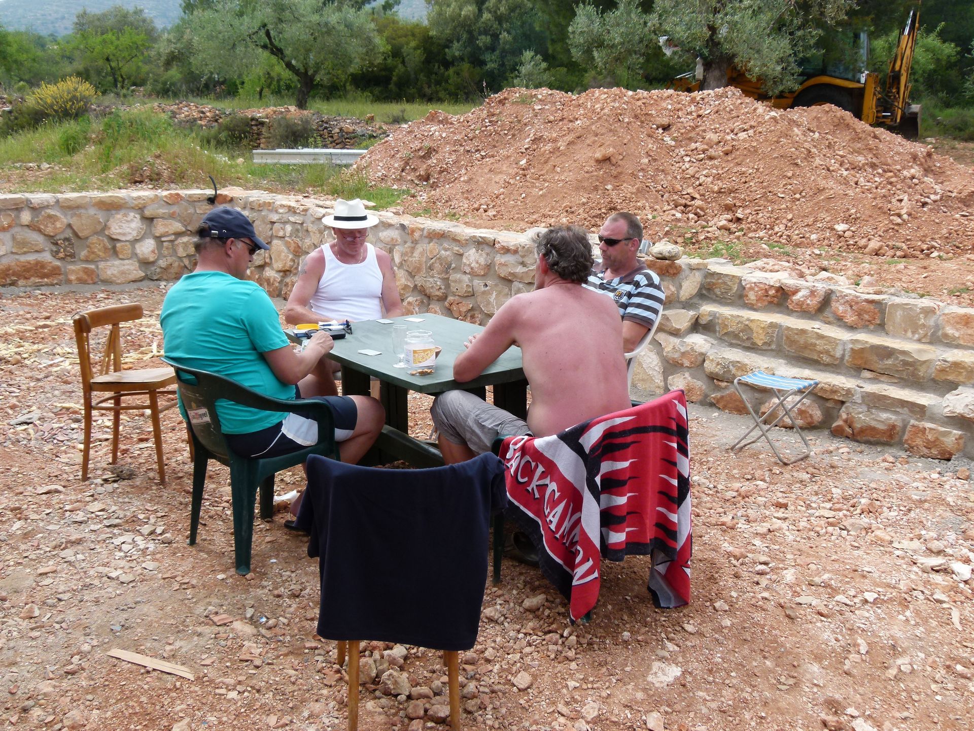 Vier mannen zitten rond een tafel en spelen een spel in de buitenlucht, met bouwwerkzaamheden op de achtergrond.