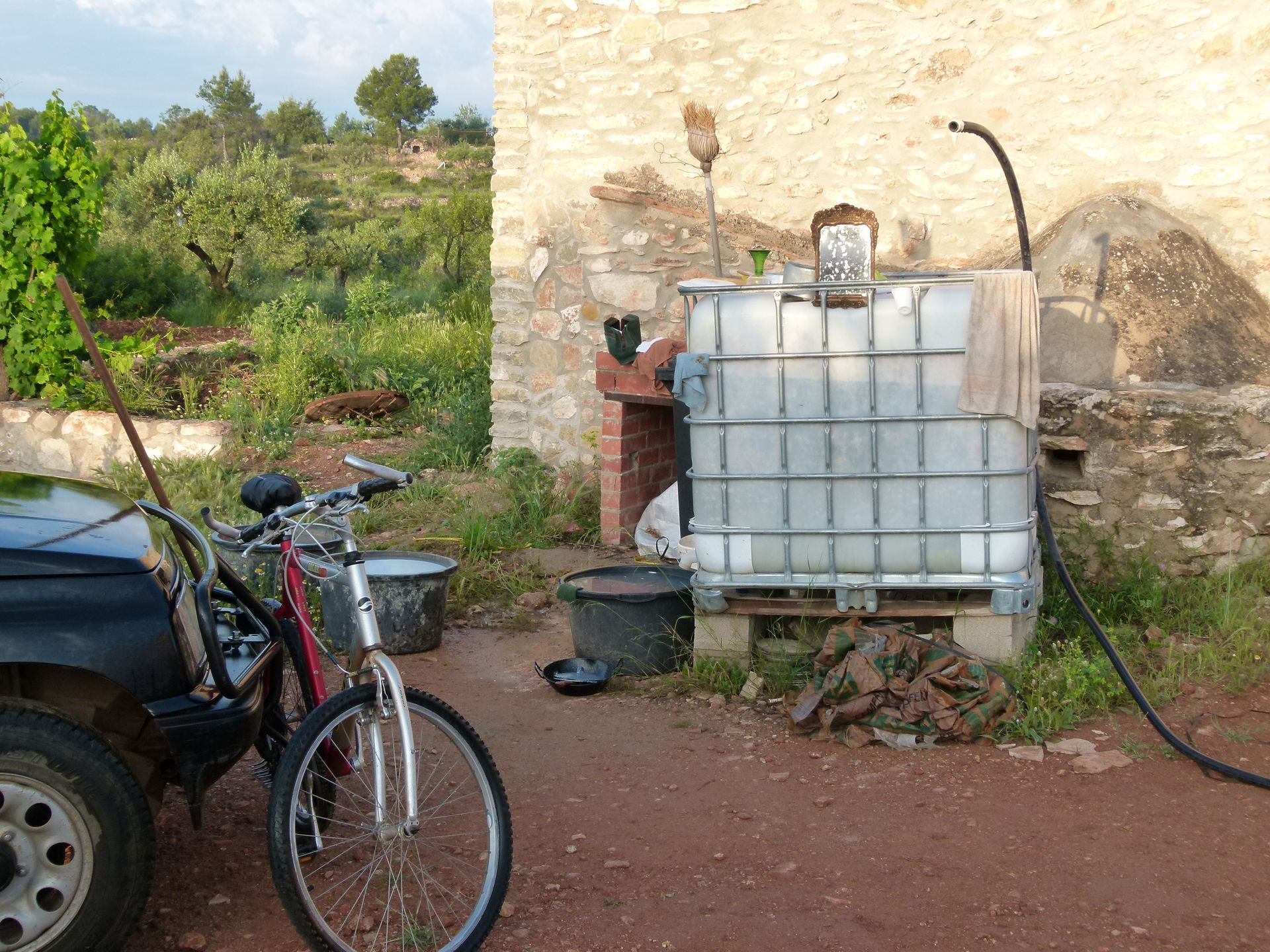 Een buitenscène met een recyclinginstallatie: IBC-tank, brandstofslang, auto, fiets en een stenen gebouw.
