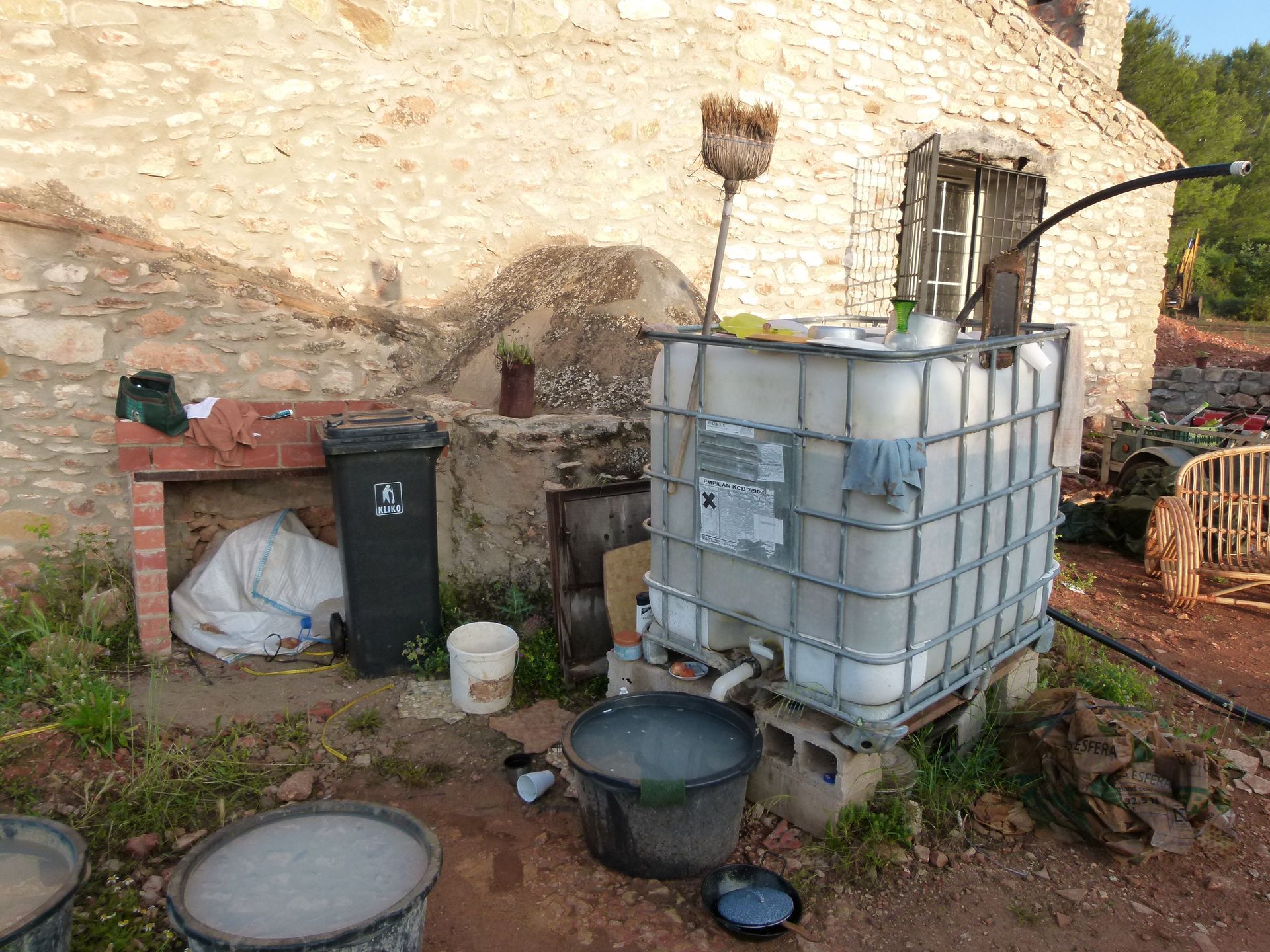 Een buitenscène: een waterreservoir op blokken met leidingen, vlakbij een bakstenen constructie en vuilnisbakken.