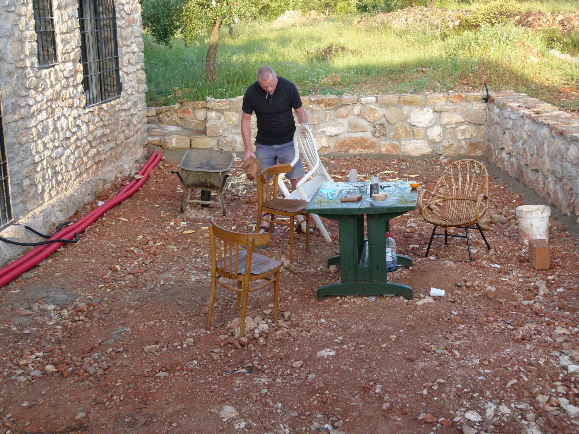 Een man staat bij stoelen en een tafel op een terras van rode aarde, naast een stenen gebouw.