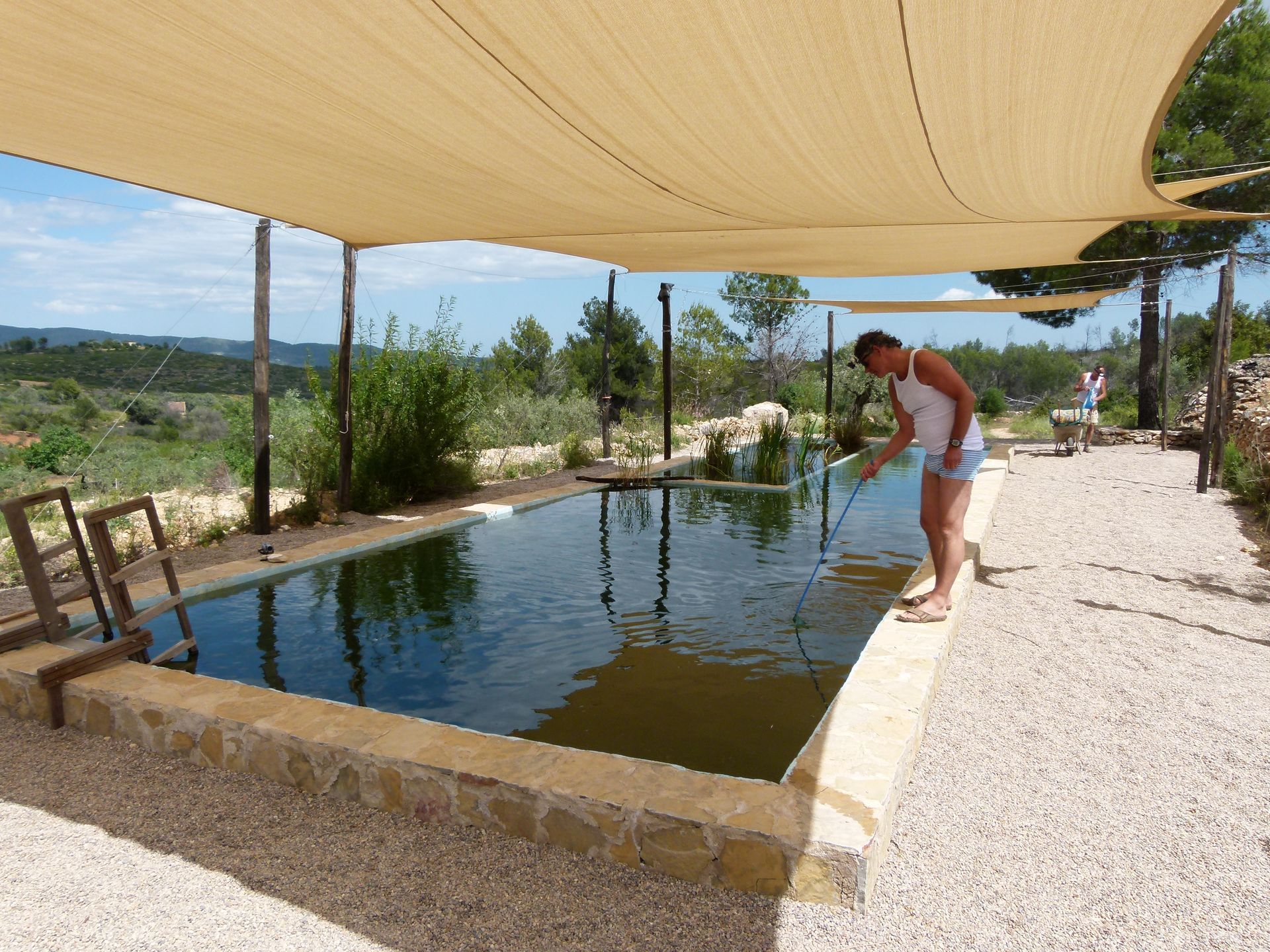 Een man onderhoudt een rechthoekig buitenzwembad onder een afdak, met een grindpad en een groen landschap in de verte.