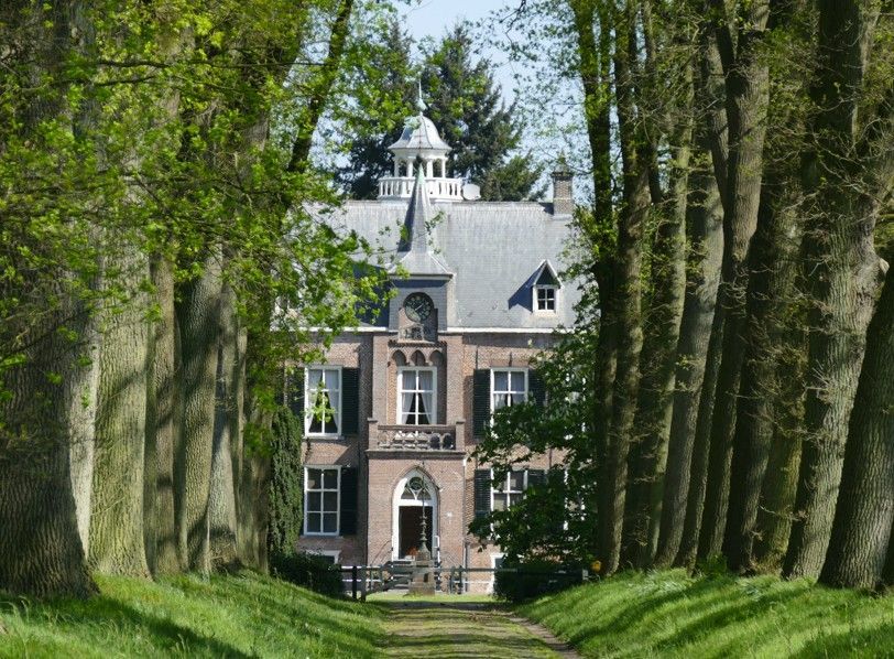 Landhuis omgeven door hoge bomen. Stenen gebouw met een grijs dak en een witte, sierlijke koepel. Zonnige buitenomgeving.