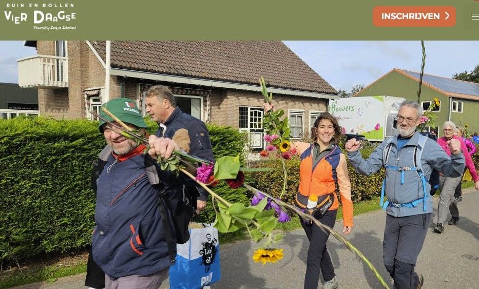 Een groep mensen loopt met bloemen, lachend, in een zonnige buitenomgeving.