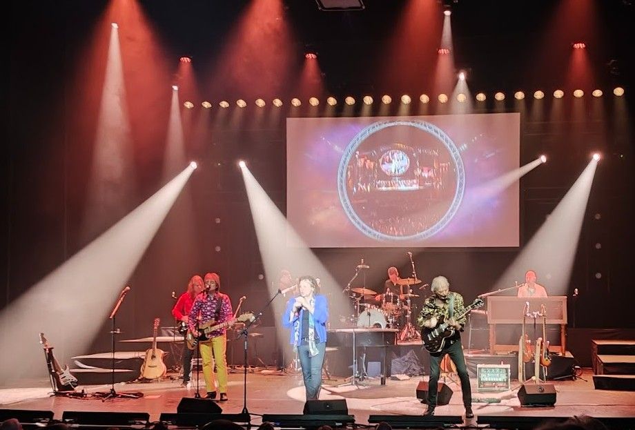 Band treedt op op een podium met felle schijnwerpers, een groot scherm als achtergrond en instrumenten.