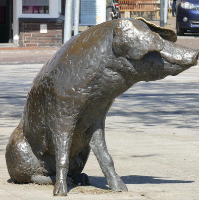 Bronzen varkensbeeldje dat op een stoep staat en naar rechts kijkt.