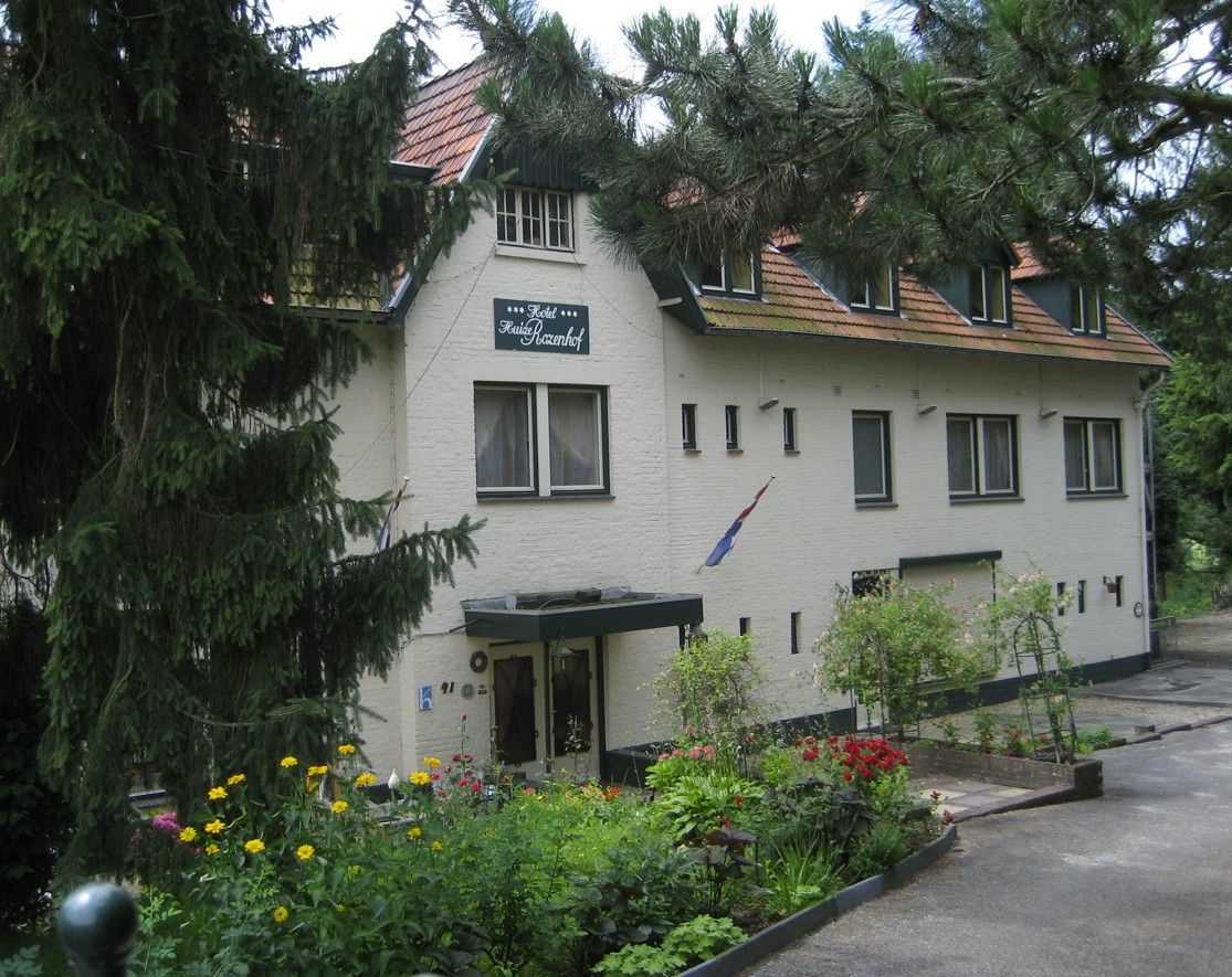 Wit, twee verdiepingen tellend huis met groene luiken, een bloementuin en bomen aan de voorzijde.