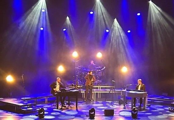 Band treedt op het podium op onder blauw-witte spotlights. De muzikanten bespelen keyboard, drums en saxofoon.
