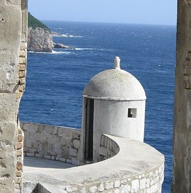Stenen toren op een vestingmuur met uitzicht op zee; blauw water en rotsachtige kust op de achtergrond.
