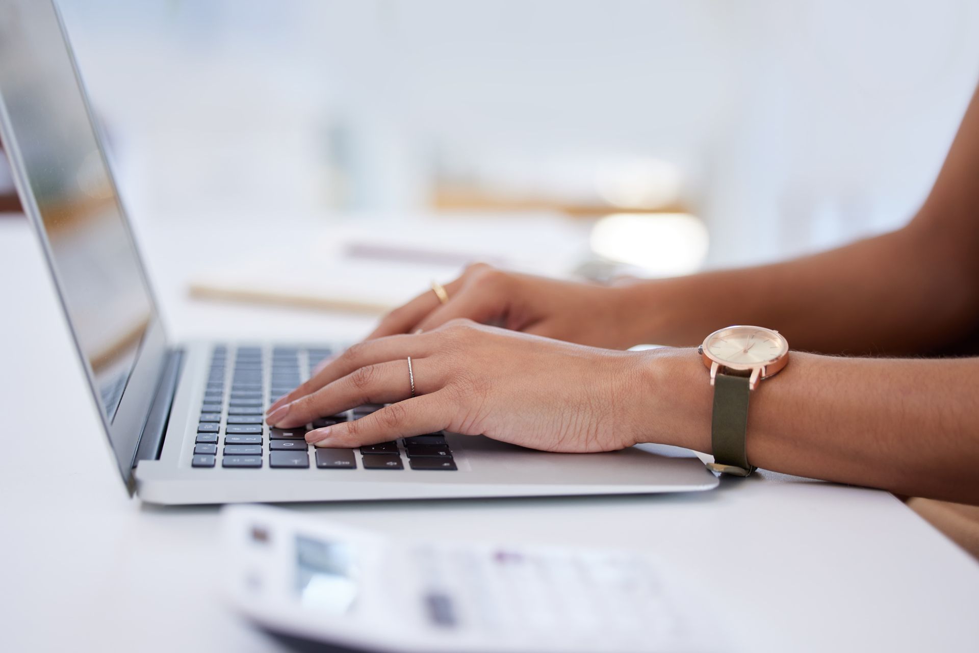 Hands typing on a laptop keyboard at a desk, wearing a wristwatch.
