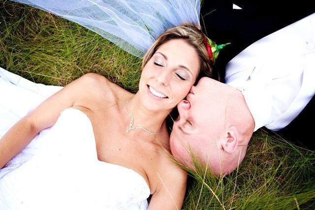 A bride and groom are laying in the grass and the bride is kissing the groom on the forehead.