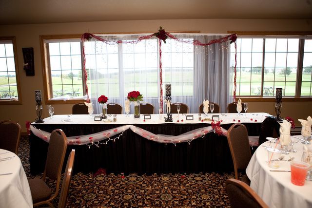 A room with tables and chairs and a long table decorated for a wedding reception.