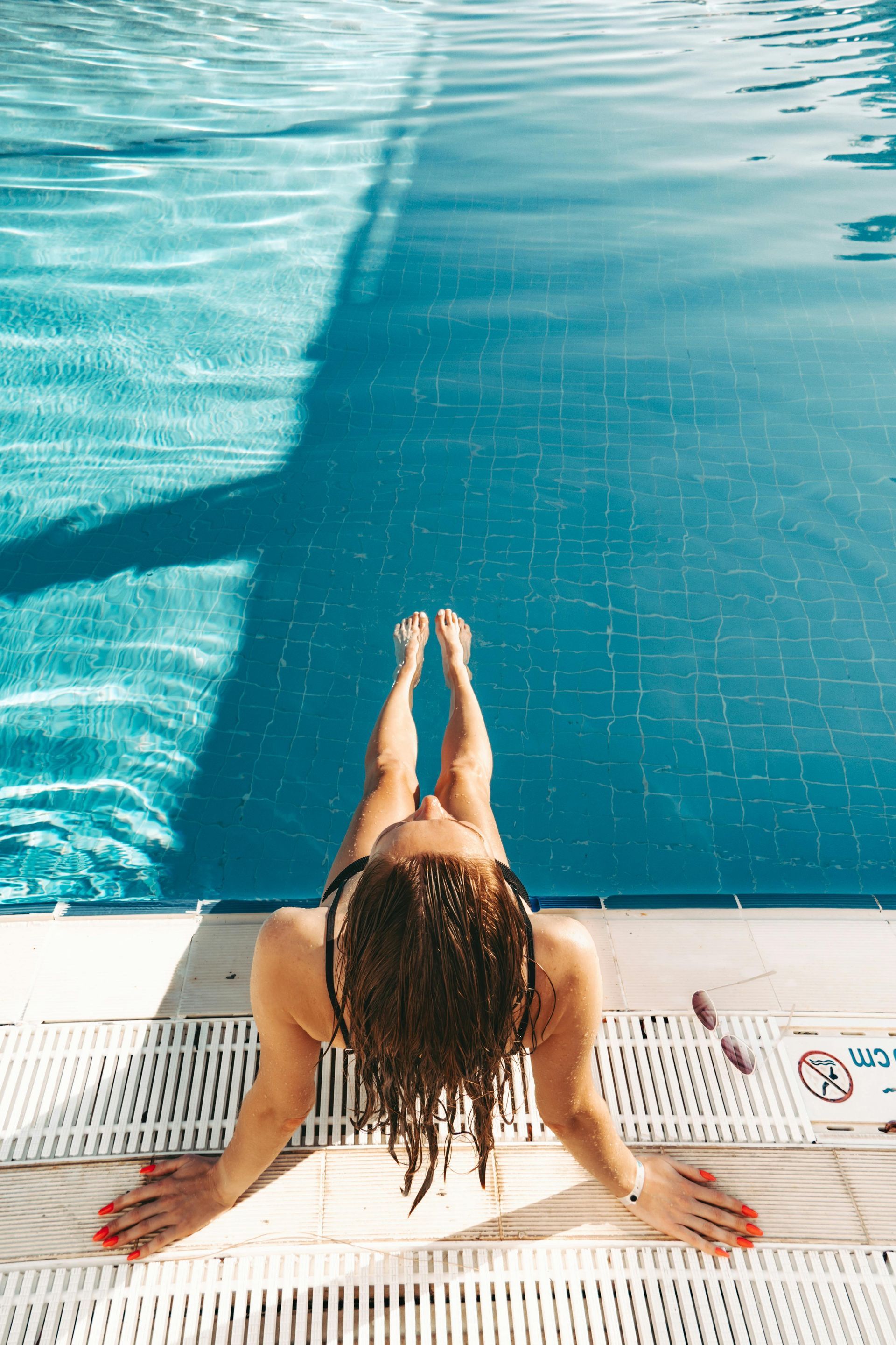 Woman sitting by a pool, legs in water. Sunlight casts a long shadow.