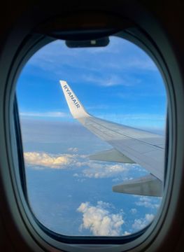 View from an airplane window: wing against blue sky, clouds below. Ryanair logo visible.