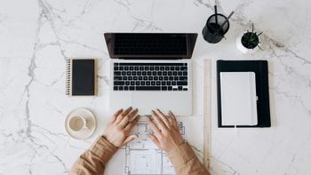 Hands working on blueprints at a marble desk with a laptop, notebook, coffee cup, and pens.