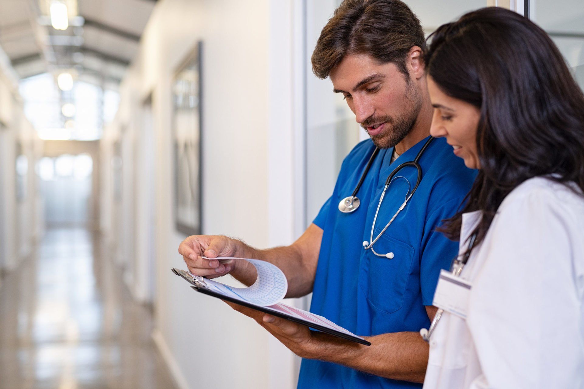 A doctor and nurse are looking at a clipboard in a hospital hallway.