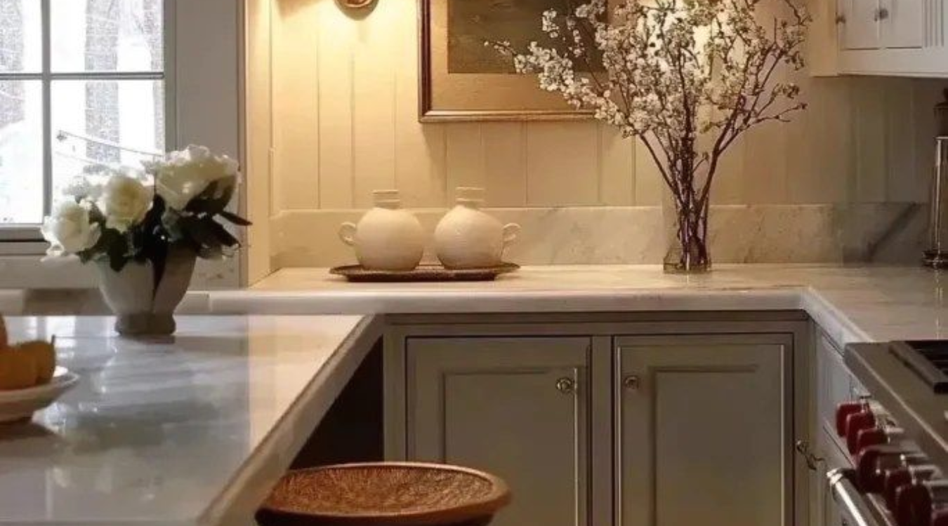 A kitchen worktop styled with white marble, light grey cabinetry, a vase of fresh flowers, two ceramic jars and a wooden stool for a soft, refined finish. A kitchen counter featuring white marble, light gray cabinets, a vase of flowers, two ceramic jars, and a wooden stool.